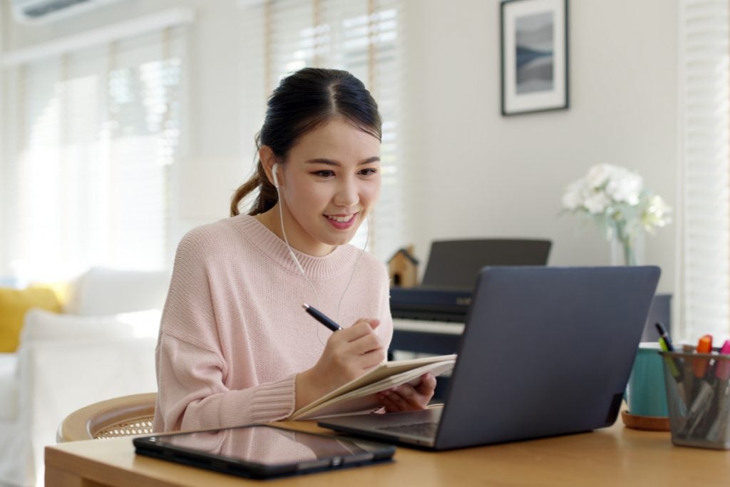 Menina de fone de ouvido estudando em frente a computador e realizando anotações